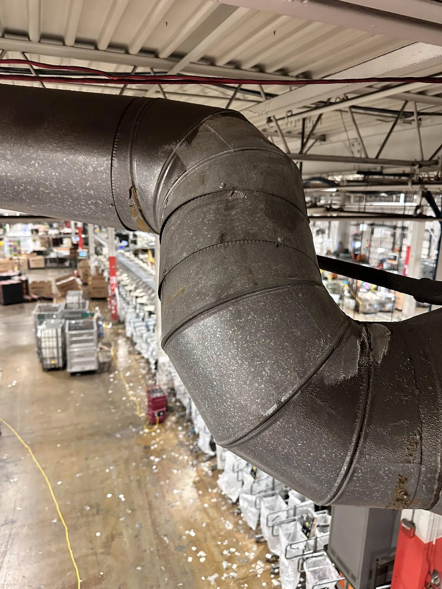 Industrial ductwork inside a large warehouse with metal pipes and visible rows of boxes and storage bins on the floor below.