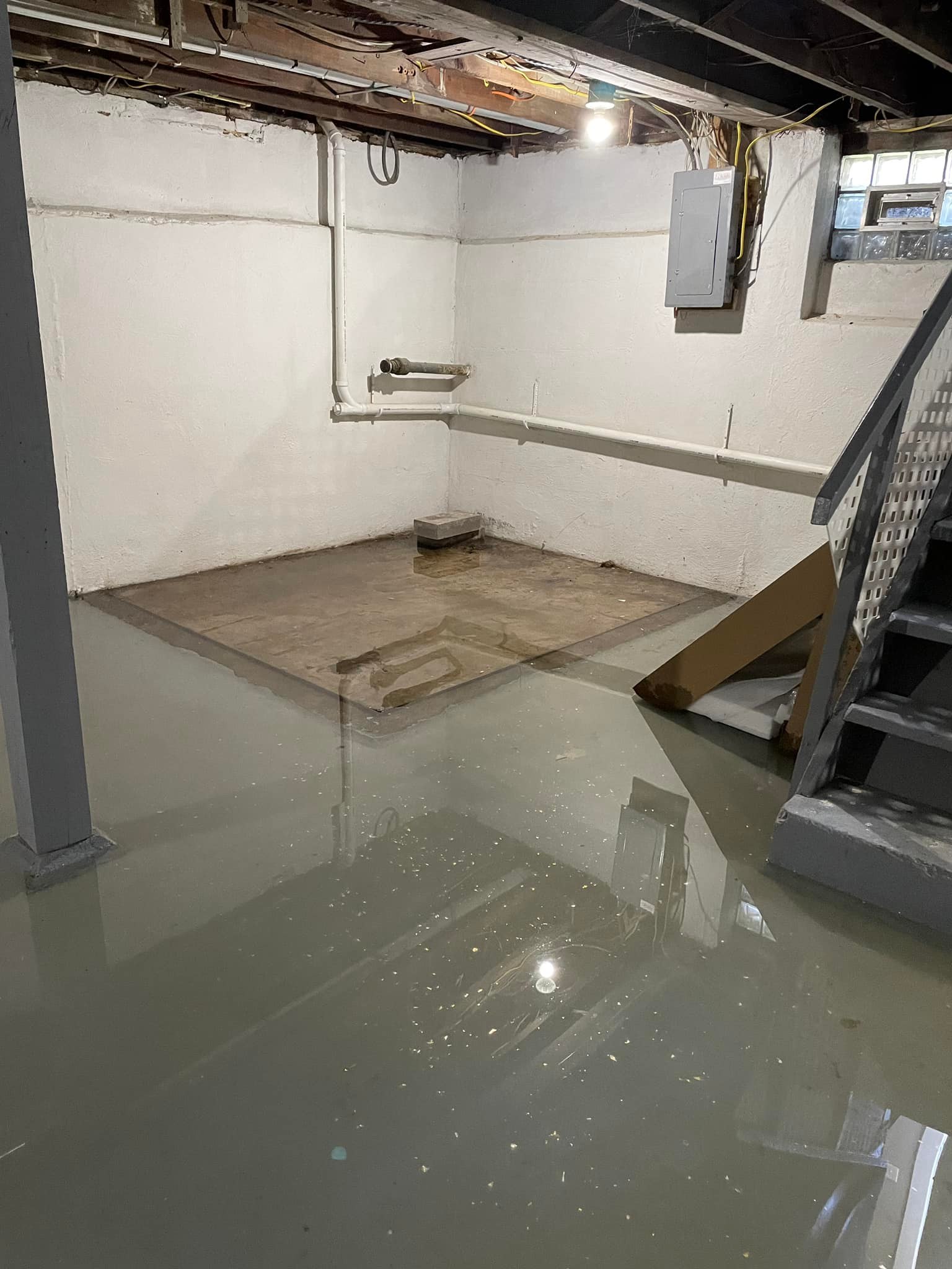 A basement with light flooding, visible water on the floor, exposed ceiling beams, and a staircase. Walls are painted white with visible pipes and an electrical panel.