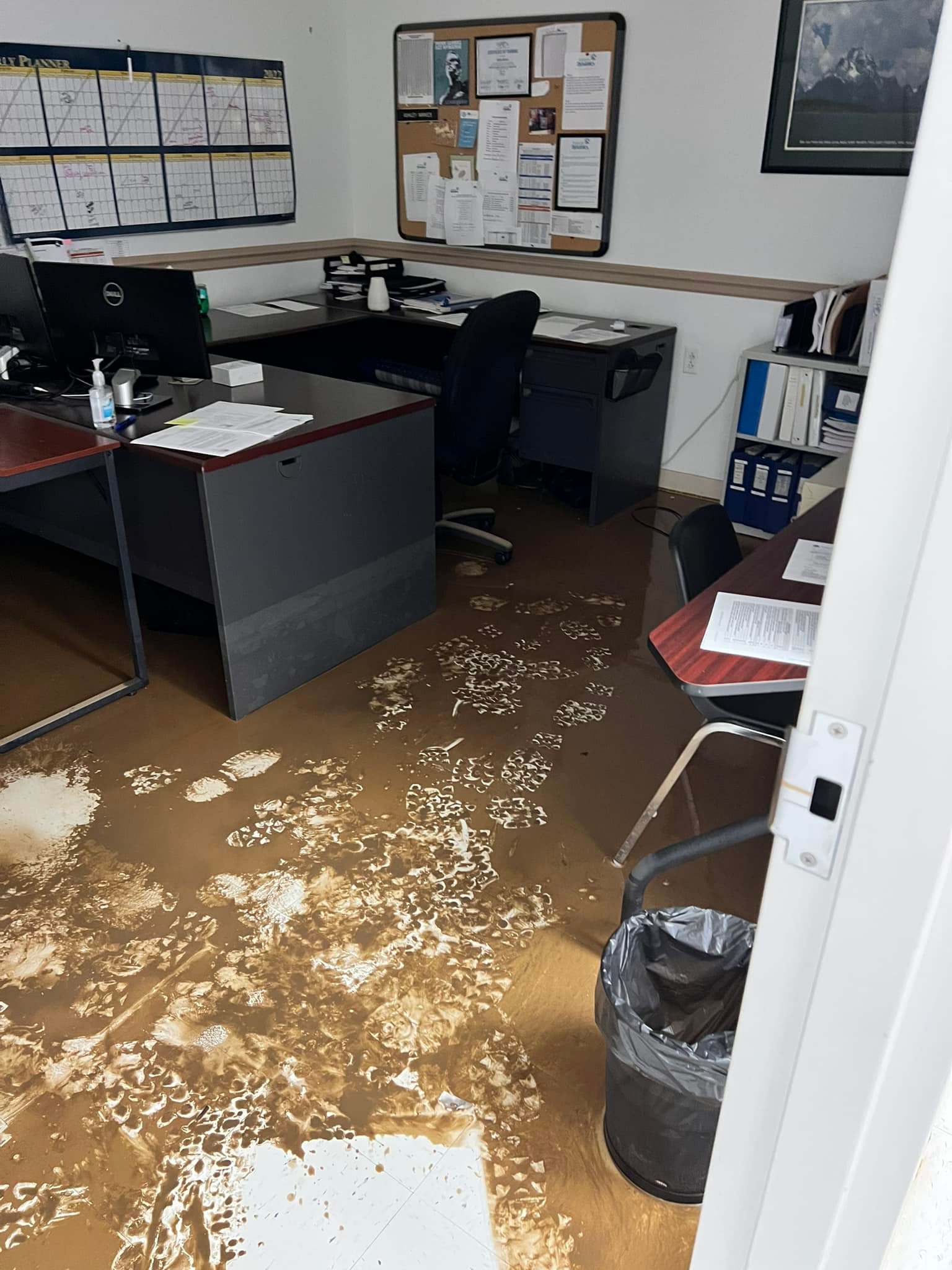 Office with two desks and chairs; floor covered in muddy footprints and water stains. Walls have a calendar and bulletin board.