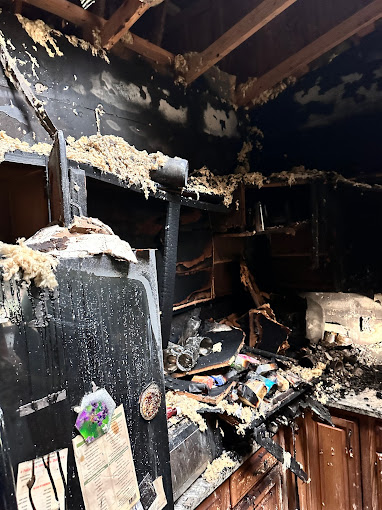 Fire-damaged kitchen with charred cabinets, a soot-covered refrigerator with magnets, and debris scattered across counters.